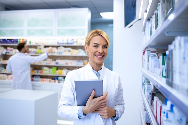 Retrato de hermosa mujer rubia farmacéutica de pie en la tienda de farmacia o droguería por medicamentos y sosteniendo tableta digital