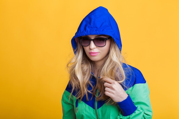 Retrato de hermosa mujer rubia con gafas de sol y chaqueta con capucha verde azul sobre fondo amarillo hola ...