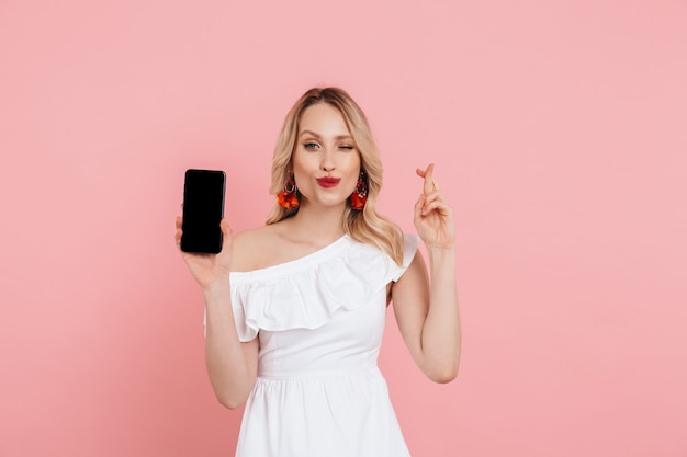 Retrato de una hermosa mujer rubia con vestido de verano que se encuentran aisladas sobre rosa, mostrando el teléfono móvil con pantalla en blanco, sosteniendo los dedos cruzados