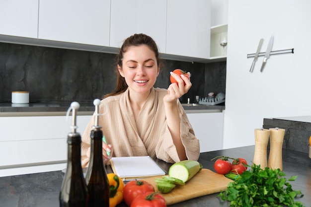 Retrato de una hermosa mujer sonriente escribiendo su menú saludable comiendo tomate mientras cocina