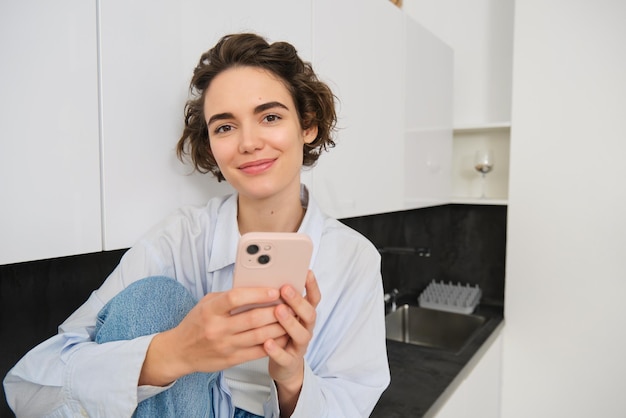 Retrato de una hermosa mujer sonriente sentada en la cocina con un teléfono inteligente mirando feliz a la cámara gastar