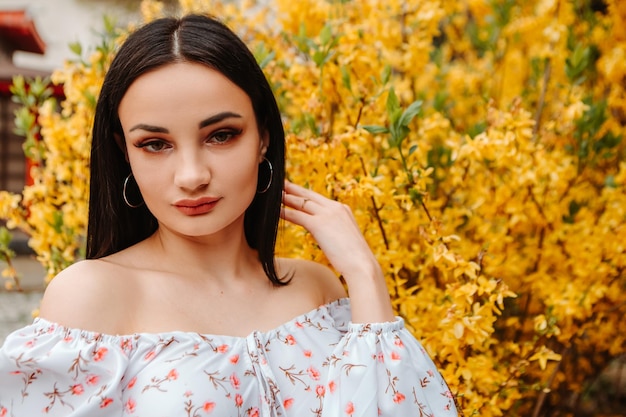 Retrato de hermosa mujer tierna vestida con un vestido florido rosa posando cerca del árbol de forsythia amarilla