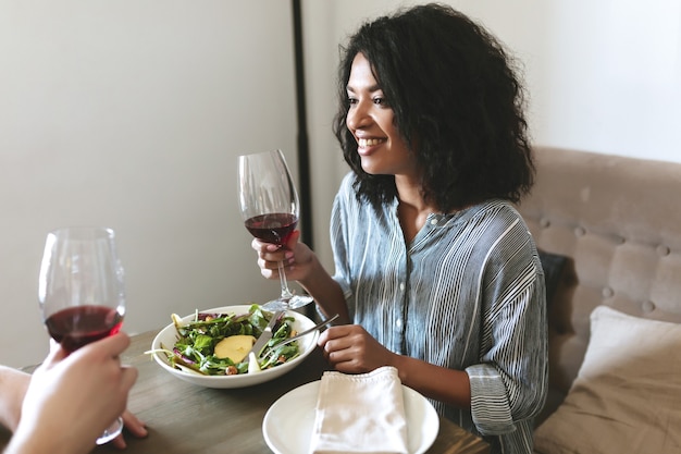 Retrato de hermosa niña afroamericana con copa de vino tinto en la mano y ensalada en la mesa en el restaurante