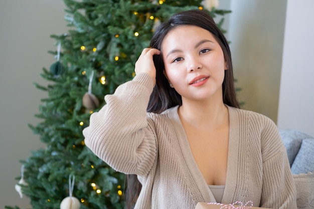 retrato de una hermosa niña asiática sonriente sentada en un sofá en casa cerca del árbol de navidad