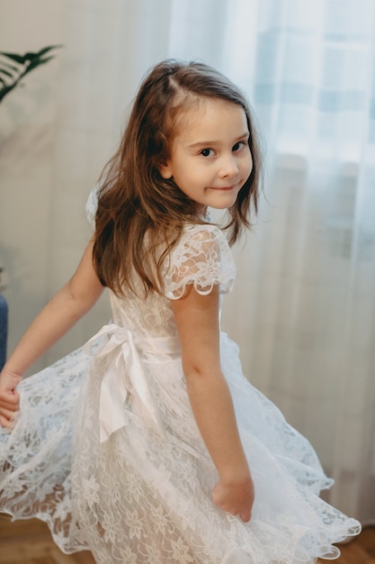 Retrato de una hermosa niña bailando vestida con un vestido blanco en casa sonriendo.