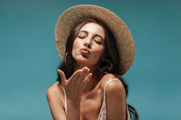 Retrato de hermosa niña con cabello largo en aretes modernos y sombrero de ala ancha fresco soplando beso con los ojos cerrados en la pared azul