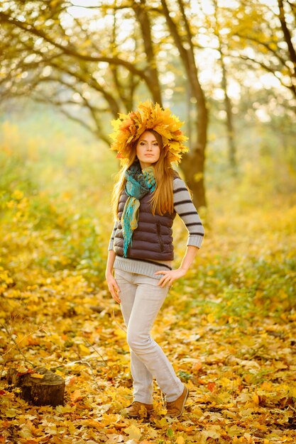 Retrato de una hermosa niña en un chaleco azul, se levanta en el contexto de un fabuloso otoño. En la cabeza hay una exuberante corona con hojas de otoño. Modelo muy tierno mirando a la cámara.