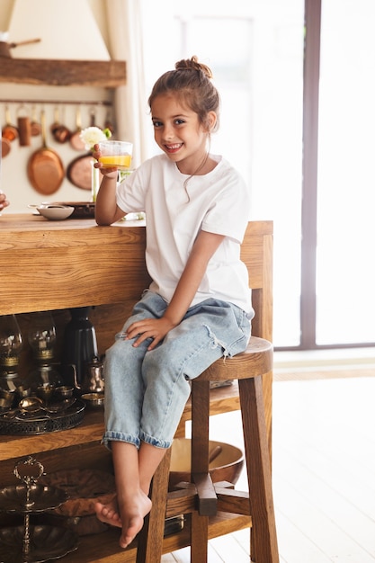 Retrato de hermosa niña desayunando y bebiendo jugo de naranja mientras está sentado a la mesa en la cocina luminosa en casa