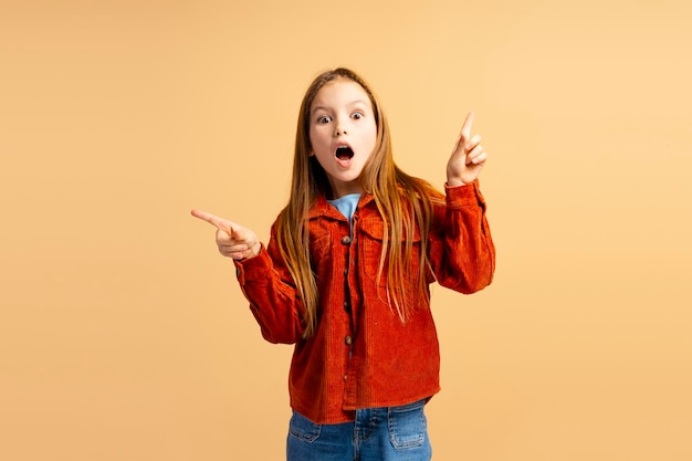 Retrato de una hermosa niña emocionada con ropa casual apuntando con los dedos al espacio de copia