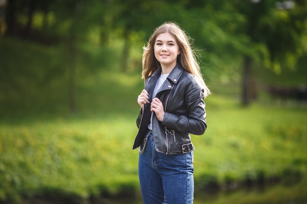 Retrato de una hermosa niña con estilo en jeans azules y chaqueta de cuero en el parque de la ciudad en el fondo del bosque verde