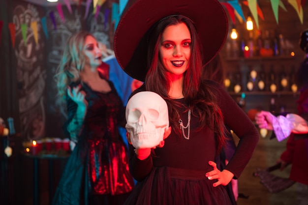 Retrato de una hermosa niña con un gran sombrero vestida como una bruja para la celebración de halloween. Bruja aterradora sosteniendo un cráneo humano mirando a la cámara