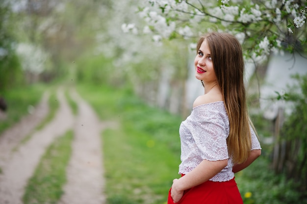 Retrato de hermosa niña con labios rojos en el jardín de flores de primavera, use vestido rojo y blusa blanca