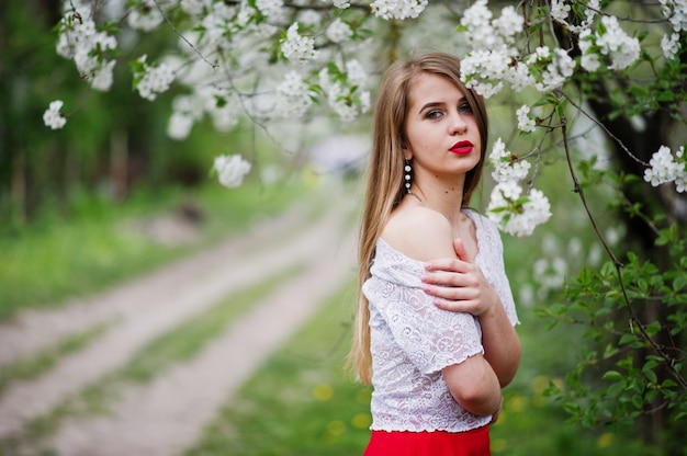 Retrato de hermosa niña con labios rojos en el jardín de flores de primavera, use vestido rojo y blusa blanca