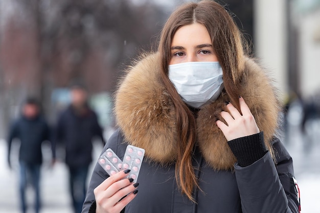 Retrato de una hermosa niña con una máscara protectora médica sosteniendo pastillas rosas en sus manos para los resfriados y la gripe Retrato de una mujer en la calle de invierno bajo la nieve