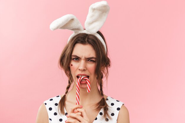 Retrato de una hermosa niña con orejas de conejo que se encuentran aisladas, haciendo muecas, sosteniendo bastón de caramelo