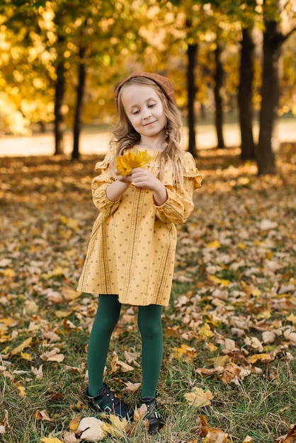 Retrato de una hermosa niña rubia con un vestido amarillo en un parque de otoño con un ramo de hojas de arce