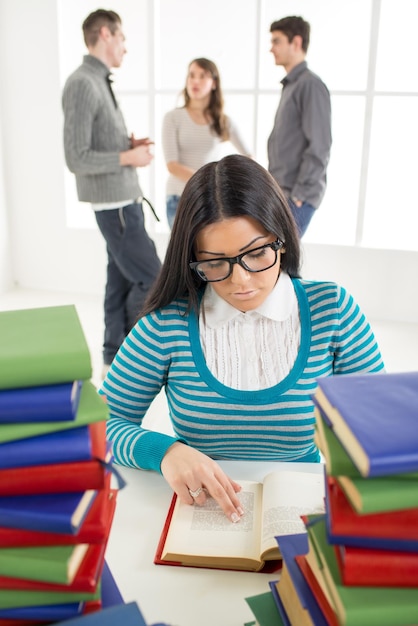 Retrato de una hermosa niña sentada y aprendiendo entre los muchos libros en primer plano. A sus amigos de pie está detrás de ella y hablando. Mirando a la cámara.