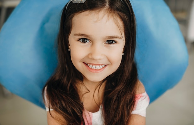 Retrato de una hermosa niña sentada en una estomatología pediátrica mirando a la cámara riendo antes de la cirugía de dientes.