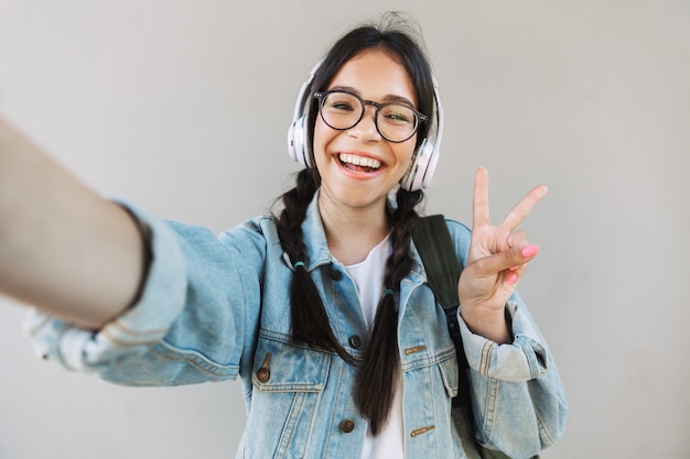 Retrato de una hermosa niña sonriente en chaqueta de mezclilla con anteojos aislados sobre pared gris escuchando música con auriculares tomar un selfie por cámara mostrando gesto de paz.