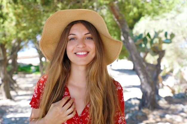 Retrato de una hermosa niña sonriente con vestido rojo y sombrero mirando a la cámara al caminar bajo los olivos