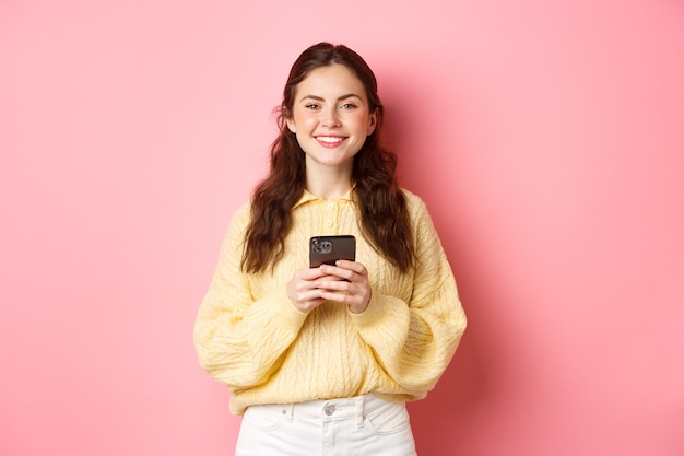 Retrato de hermosa niña sosteniendo el teléfono móvil y sonriendo, comprando en línea, usando la aplicación de redes sociales, de pie contra la pared rosa en ropa casual.