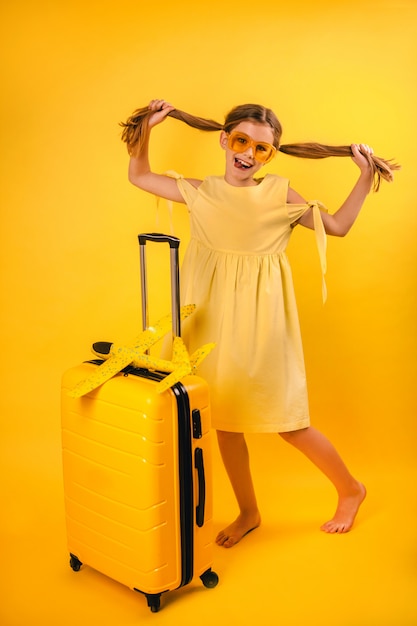 Retrato de una hermosa niña en un vestido amarillo con coletas de pie cerca de una maleta