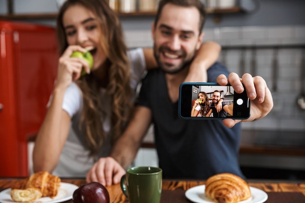 Foto retrato de hermosa pareja hombre y mujer tomando foto selfie en teléfono celular mientras desayuna en la cocina de casa