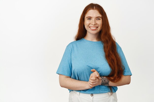 Retrato de una hermosa pelirroja tomándose de la mano, sonriendo complacida, lista para ayudar, ofreciendo su consulta, de pie con una camiseta azul sobre fondo blanco. copia espacio