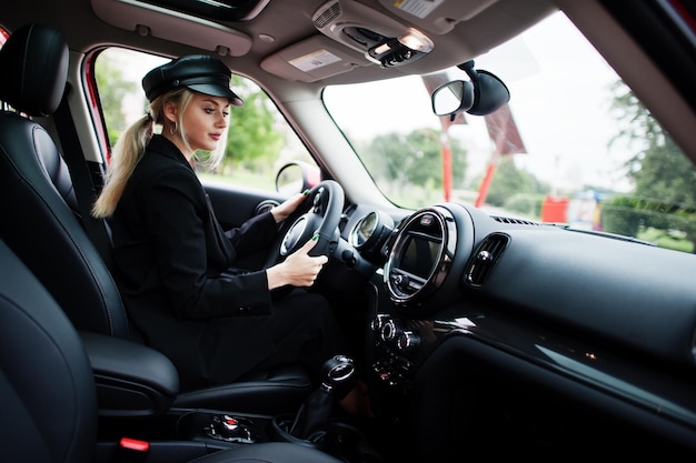Retrato de hermosa rubia sexy modelo de mujer de moda en gorra y en todo negro con maquillaje brillante sentarse y conducir coche rojo de la ciudad