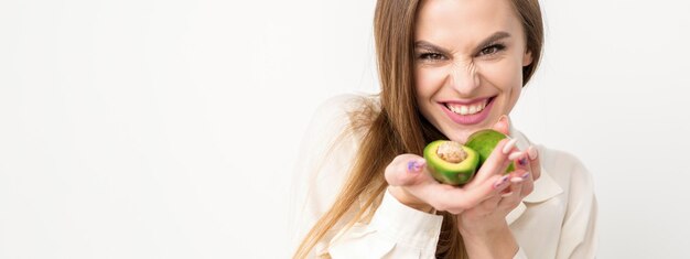 Retrato de una hermosa y sonriente joven morena caucásica con la camisa blanca con el pelo largo sosteniendo y mostrando aguacate de pie aislado sobre fondo blanco