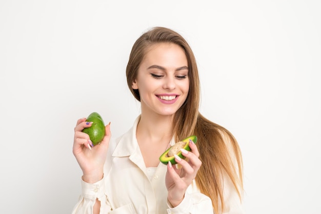 Retrato de una hermosa y sonriente joven morena caucásica con la camisa blanca con el pelo largo sosteniendo y mostrando aguacate de pie aislado sobre fondo blanco