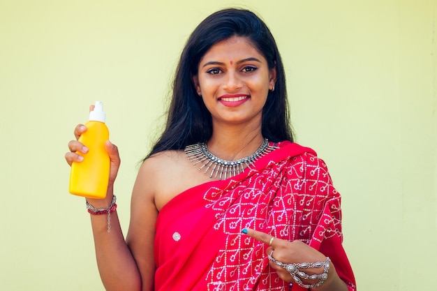 Retrato de una hermosa y sonriente sonrisa blanca como la nieve mujer india cabello negro y piel oscura en un sari rojo tradicional sosteniendo una botella de spray de protección solar en una chica de playa disfrutando de unas vacaciones en el paraíso del cuerpo spf