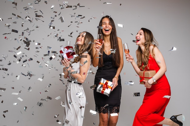 Retrato de hermosas damas joviales despreocupadas en vestidos de cóctel con vasos de bebidas alcohólicas bailando y divirtiéndose bajo la caída de confeti plateado