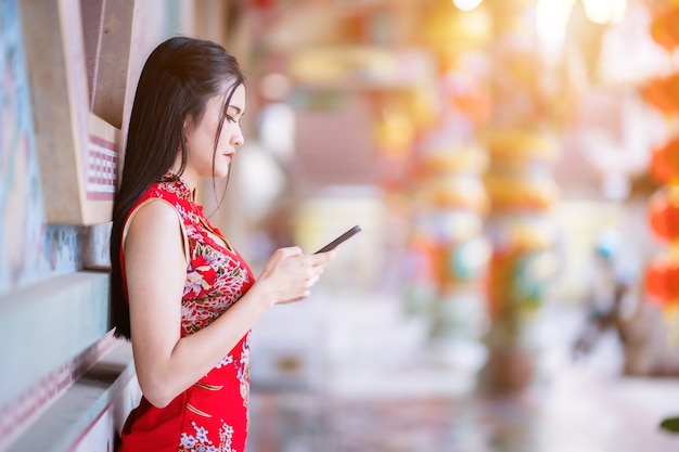 Retrato hermosas sonrisas Mujer joven asiática con cheongsam chino tradicional rojo y mensaje de escritura en el teléfono inteligente para el Festival del Año Nuevo Chino en el santuario chino