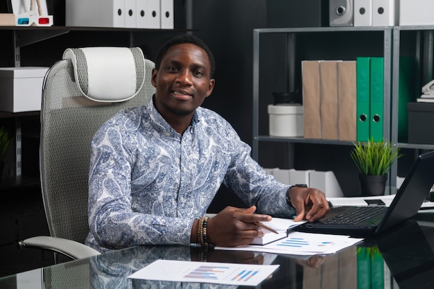 Retrato de hermoso joven empresario afroamericano que trabaja con documentos y computadora portátil en la oficina