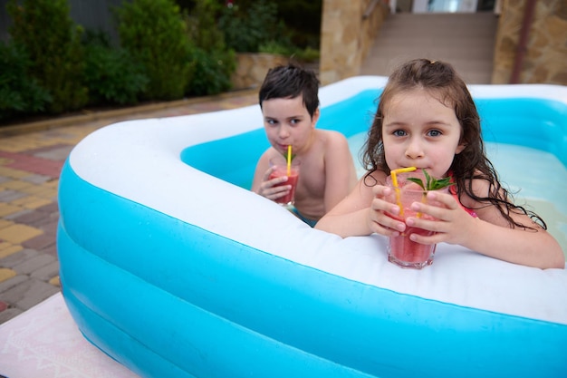 Retrato de hermosos hermanos bebiendo cócteles y divirtiéndose en la piscina al aire libre en el jardín trasero de la casa