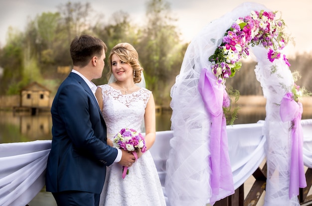 Retrato de hermosos recién casados sonrientes posando en el arco floral al atardecer