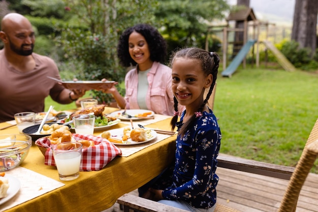 Retrato de una hija biracial feliz en la mesa comiendo con sus padres en el jardín. Verano, familia, unión, comida y estilo de vida, sin alteraciones.