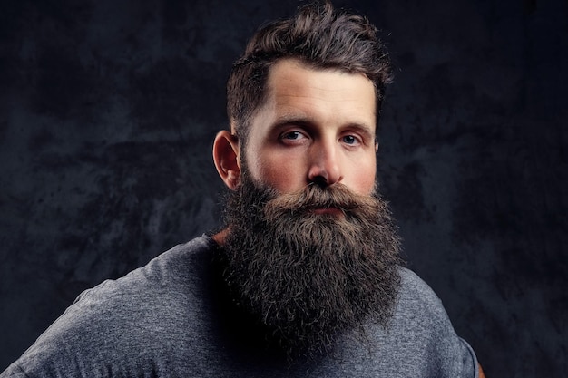 Retrato de un hipster con barba completa y corte de pelo elegante, vestido con una camiseta gris, mirando a la cámara, se encuentra en un estudio sobre un fondo oscuro.