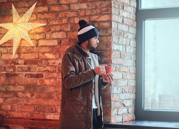 Retrato de un hipster indio barbudo vestido con una chaqueta cálida y un sombrero, bebe café de una taza roja cerca de la pared de un ladrillo rojo.