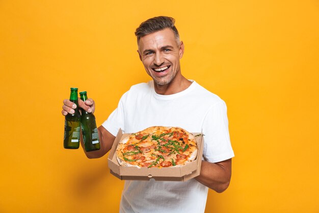 Retrato de hombre adulto de 30 años en camiseta blanca bebiendo cerveza y comiendo pizza mientras está de pie aislado en amarillo