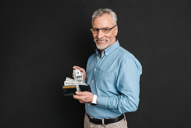 Retrato de hombre adulto 60s con canas y barba sonriendo y sosteniendo la billetera llena de billetes de un dólar, aislado sobre la pared negra