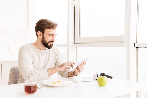 Retrato de hombre adulto sano en camisa casual sonriendo y desayunando vegetariano en el apartamento, mientras usa la tableta