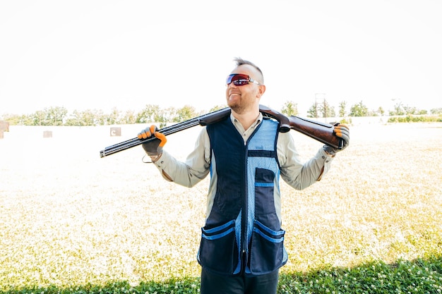 Retrato de un hombre adulto sonriente con gafas de sol, auriculares protectores y un chaleco de rifle con escopeta