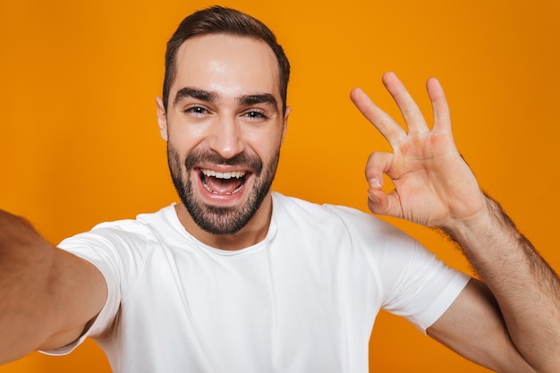 Retrato de hombre sin afeitar en camiseta sonriendo mientras toma una foto selfie, aislado en amarillo