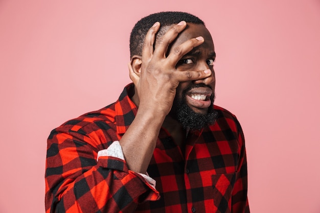 Retrato de un hombre africano asustado vestido con camisa a cuadros que se encuentran aisladas sobre la pared rosa, haciendo muecas