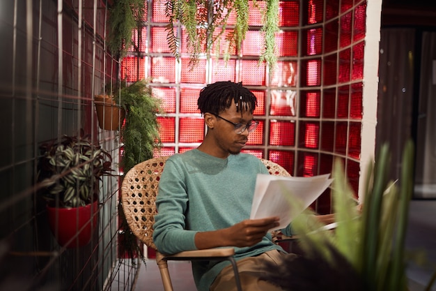 Retrato de hombre africano contemporáneo que trabaja con documentos mientras está sentado en el salón de café decorado con plantas