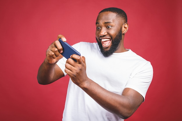 El retrato de un hombre africano joven alegre se vistió en juegos casuales en el teléfono móvil aislado sobre fondo rojo.