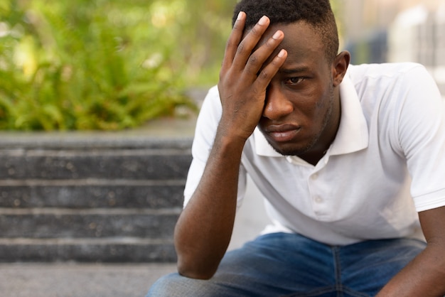 Retrato de hombre africano joven estresado mirando deprimido mientras está sentado al aire libre