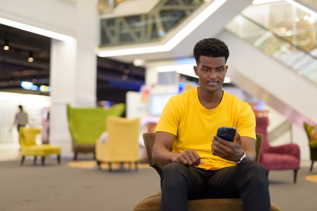 Retrato de un hombre africano negro guapo con camiseta amarilla mientras está sentado y usando el teléfono móvil